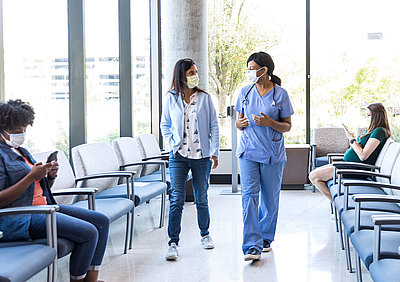 Highest LANCOM cybersecurity for inpatient clinics and hospitals Young doctor in light blue jumpsuit picks up patient with mouth guard from bright waiting area of a clinic for her treatment, where a pregnant woman and another woman are waiting to be called up